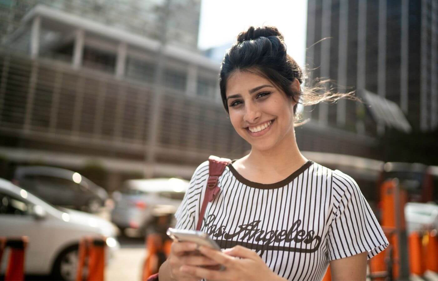 Mulher segurando um celular enquanto sorri. E segundo plano está uma rua com carros e alguns prédios desfocados